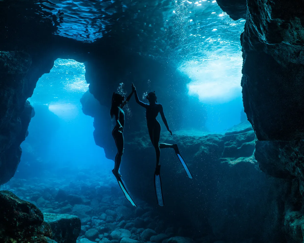 Two freedivers reaching for each other’s hands in a sunlit underwater cave